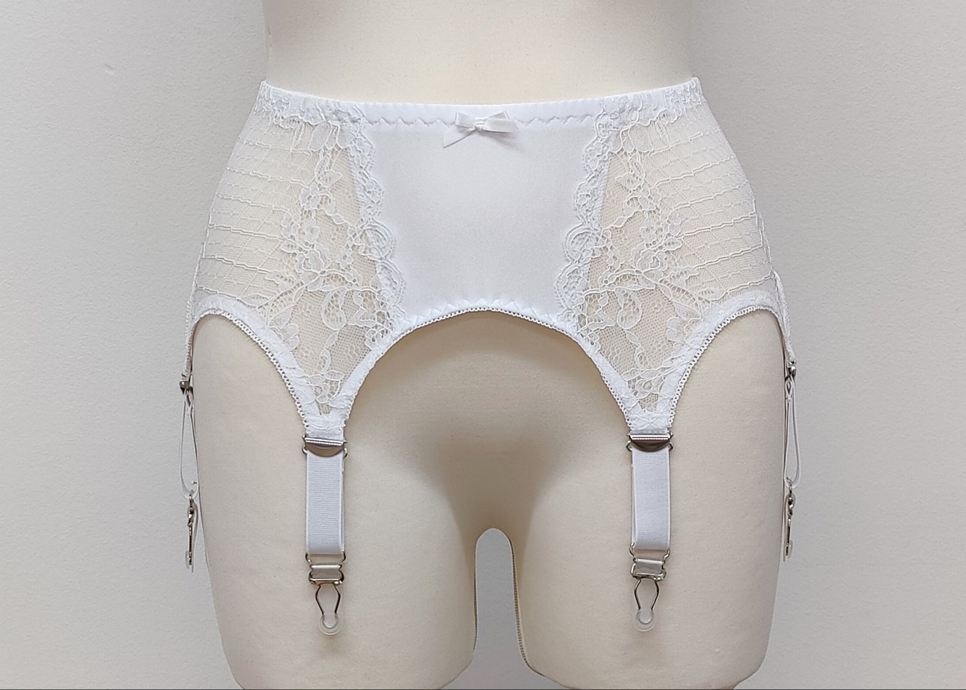 A white lace Mona garter belt with 6 adjustable straps and hook and eye closure, displayed on a mannequin.
