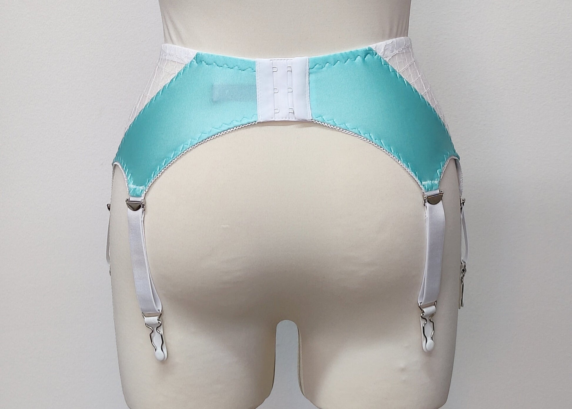 A white lace and Aqua blue Lycra, Mona garter belt with 6 adjustable straps and hook and eye closure, displayed on a mannequin, back view