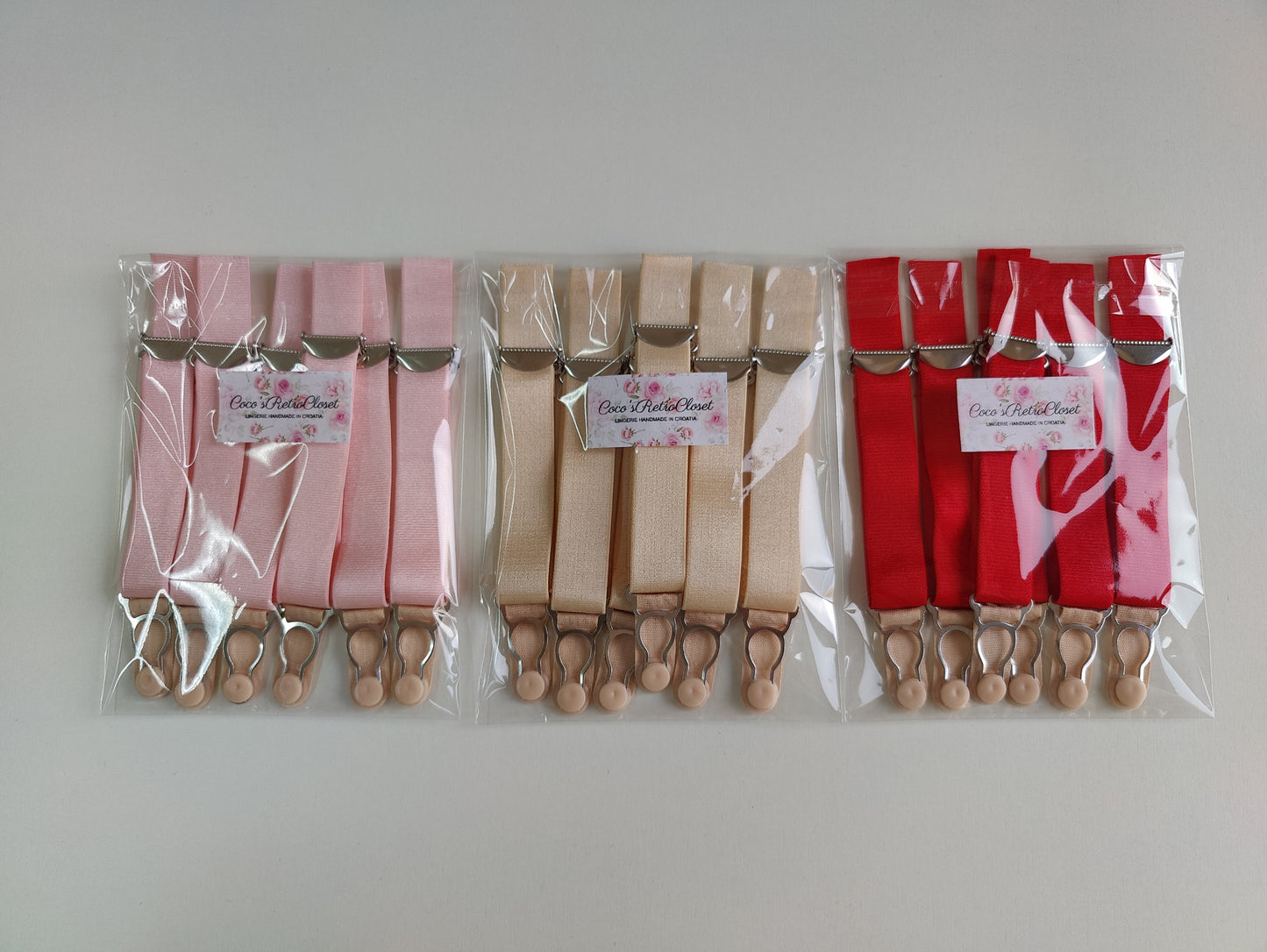 Three packages of suspenders in pink, beige, and red, each containing six straps with beige grips, sealed in clear plastic bags with white brand labels.