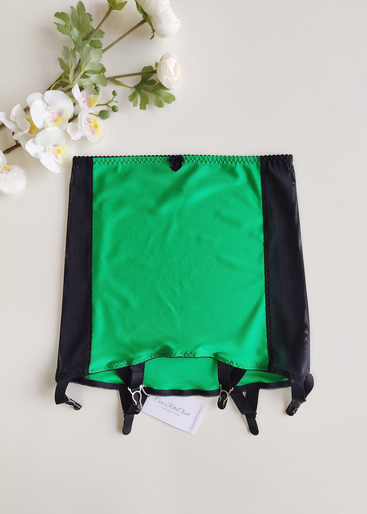 A green and black garter belt laid flat on a light background . The belt features a vibrant emerald green center panel, black side panels, black straps